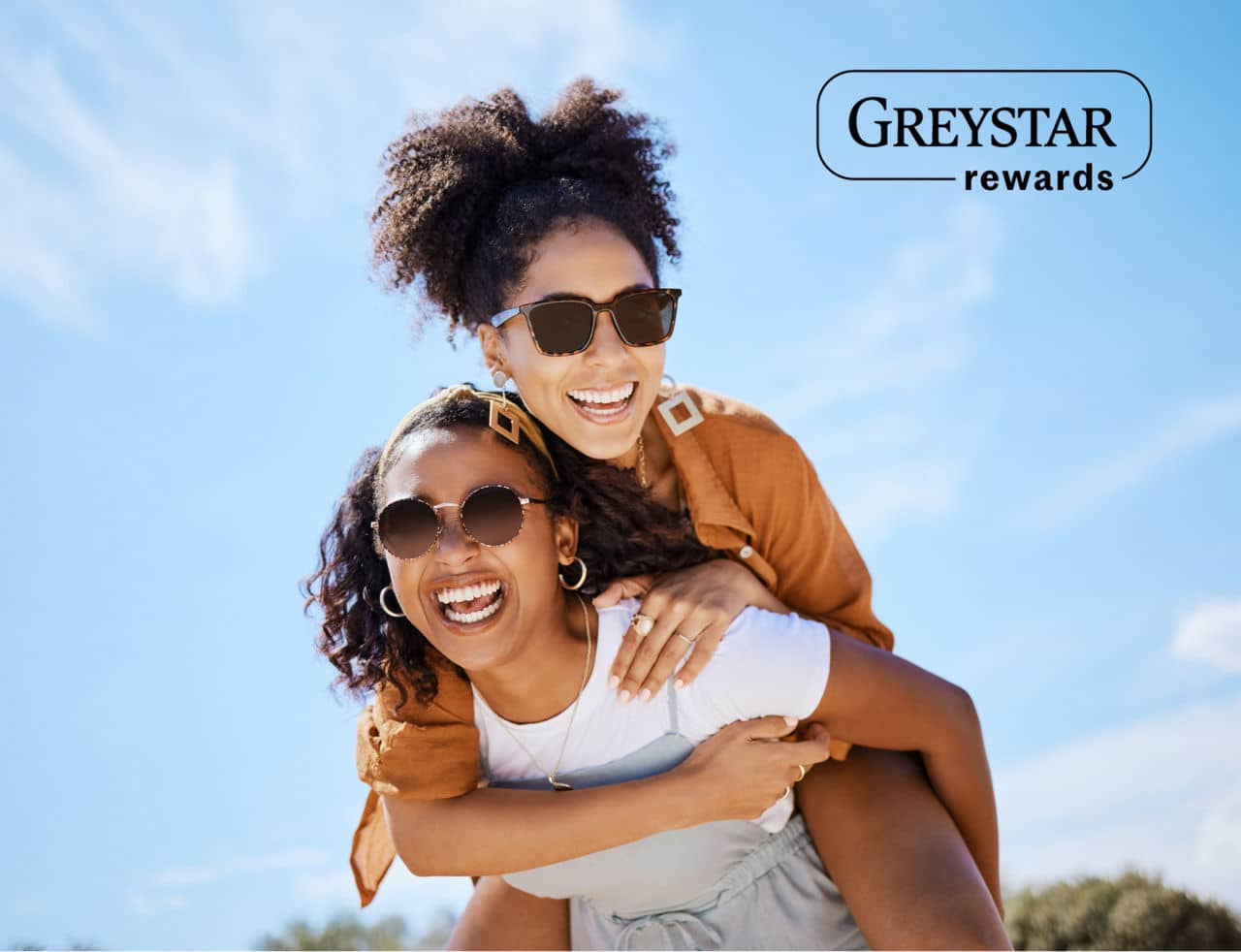 Two women outside, one carrying the other on her back while laughing and smiling with a blue sky in the background and Greystar rewards logo in the top left of the image.