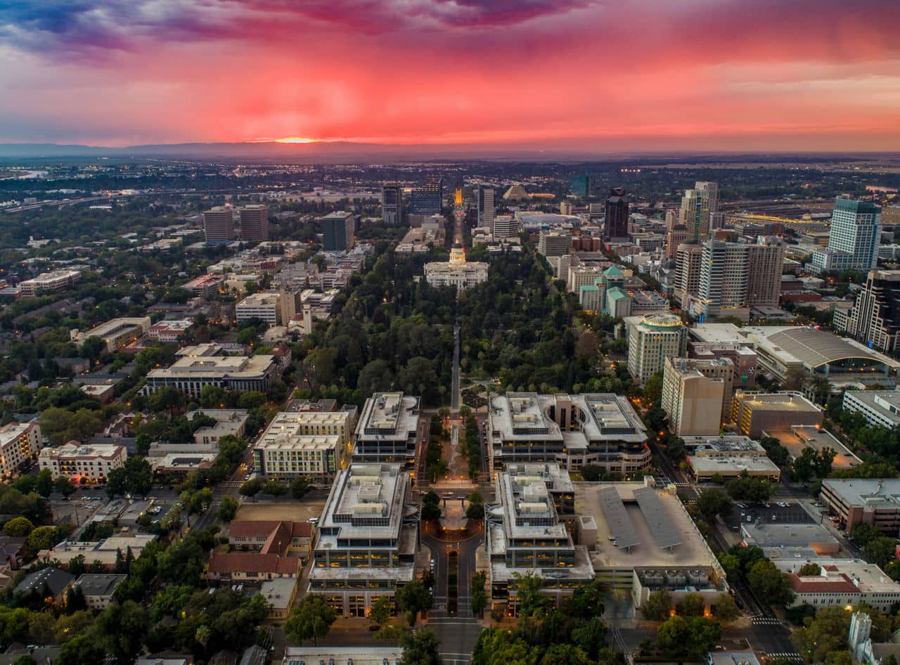 Aerial view of Sacramento at sunset, highlighting the city&apos;s skyline, the Tower Bridge, and the winding Sacramento River with a golden glow in the sky.