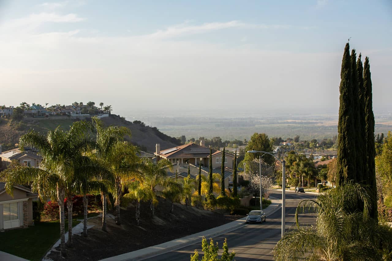A quiet suburban street in Corona, California, lined with palm trees and houses, with distant hills and open land in the background.