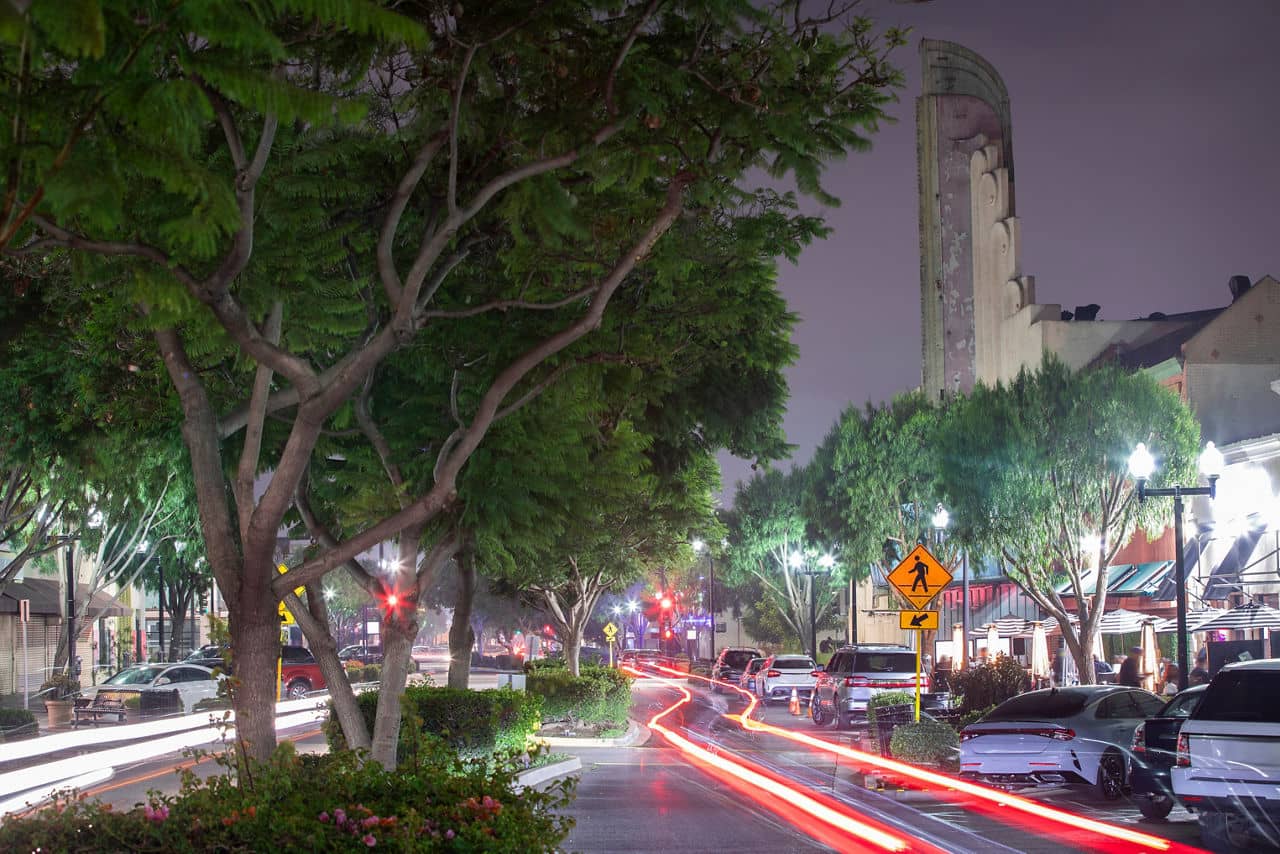A lively street in Inglewood, California, at night with glowing streetlights, trees lining the road, and light trails from passing cars.