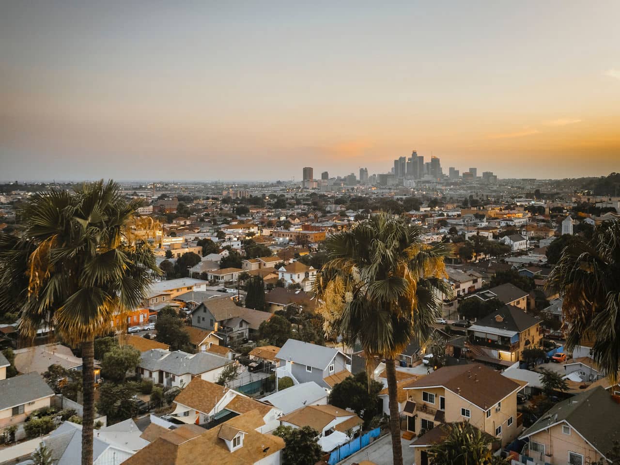 A sunset view of Lincoln Heights, California, with palm trees, houses, and a distant city skyline under a soft orange sky.