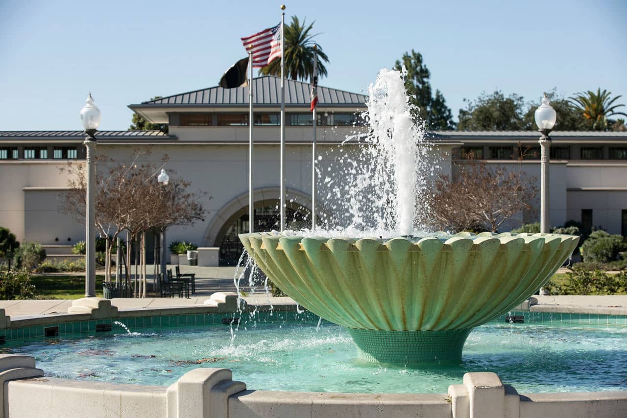 Fountain in Monrovia, California, with a flowing water feature, flagpoles, and a civic building framed by trees in the background.