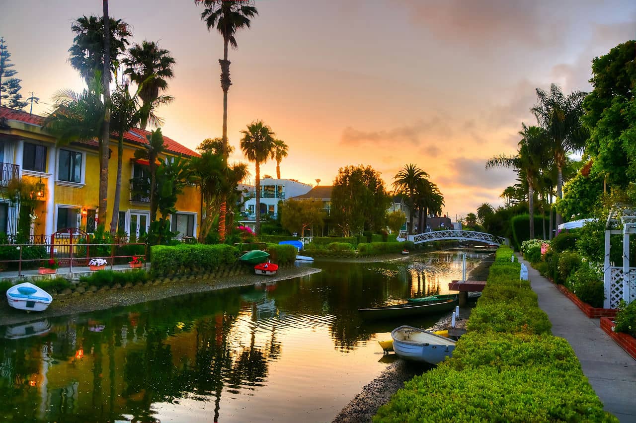 Sunset view of peaceful canals lined with colorful homes and palm trees in Venice Beach, California, with small boats docked along the water.