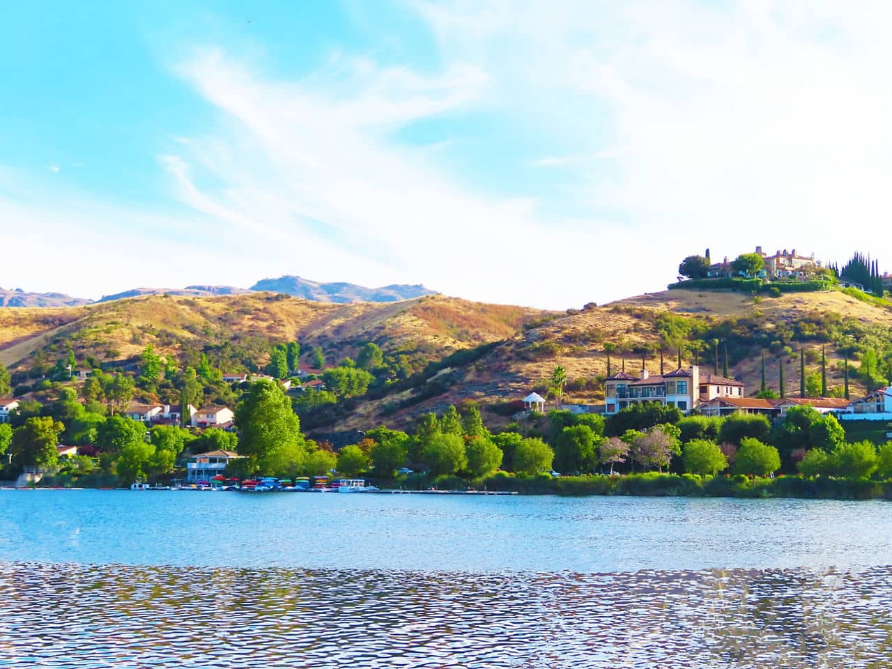A peaceful lake with hillside homes surrounded by greenery in Westlake, California, under a bright blue sky with wispy clouds.