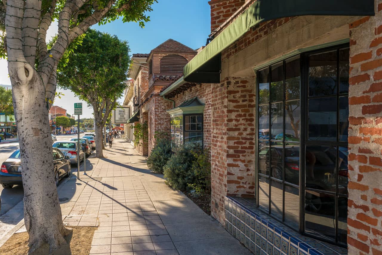 A sunny street in Westwood, California, lined with brick buildings, green awnings, parked cars, and trees providing shade on the sidewalk.