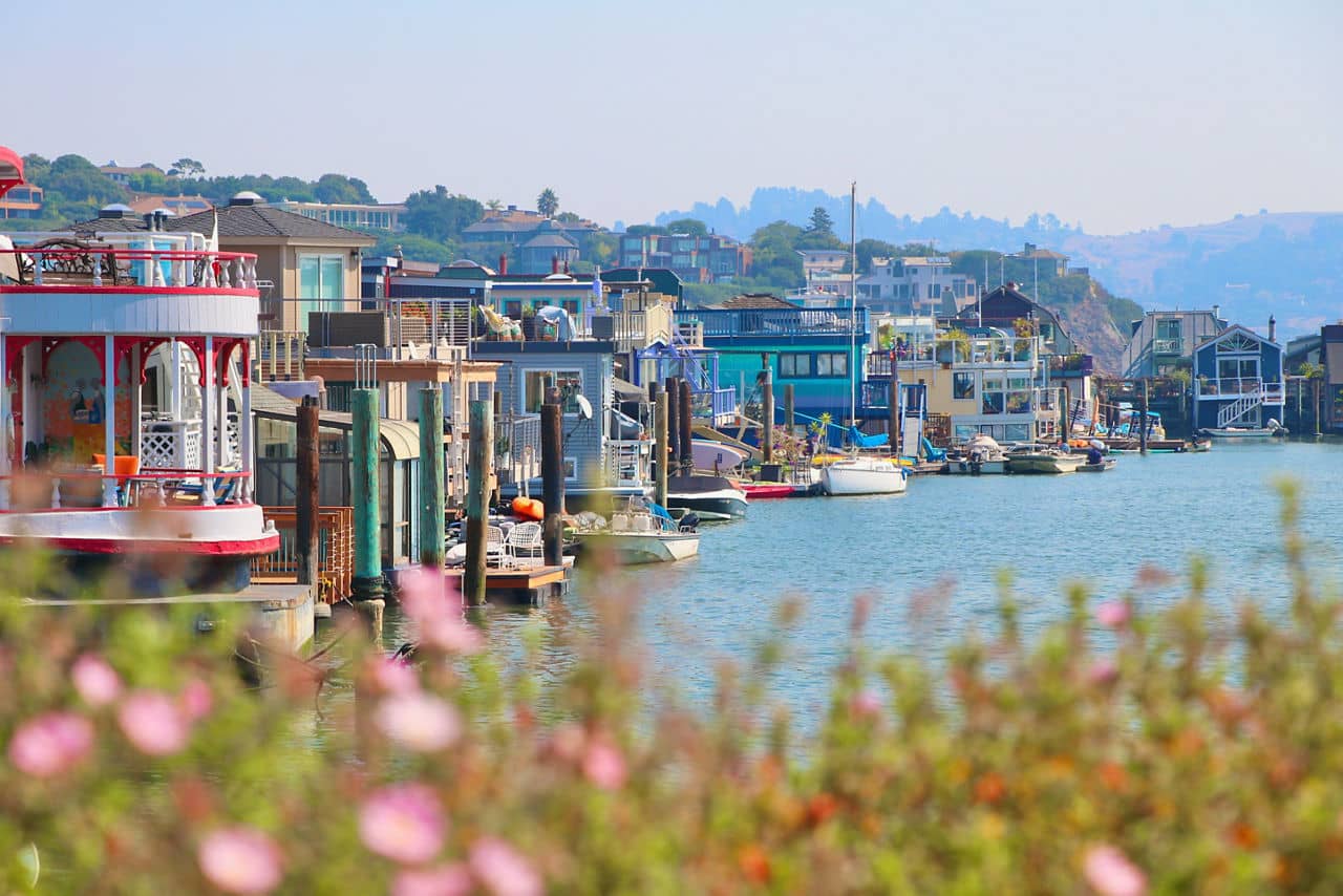 Colorful houseboats line a calm waterfront in Larkspur, California, with boats docked and hills in the background under a hazy sky.