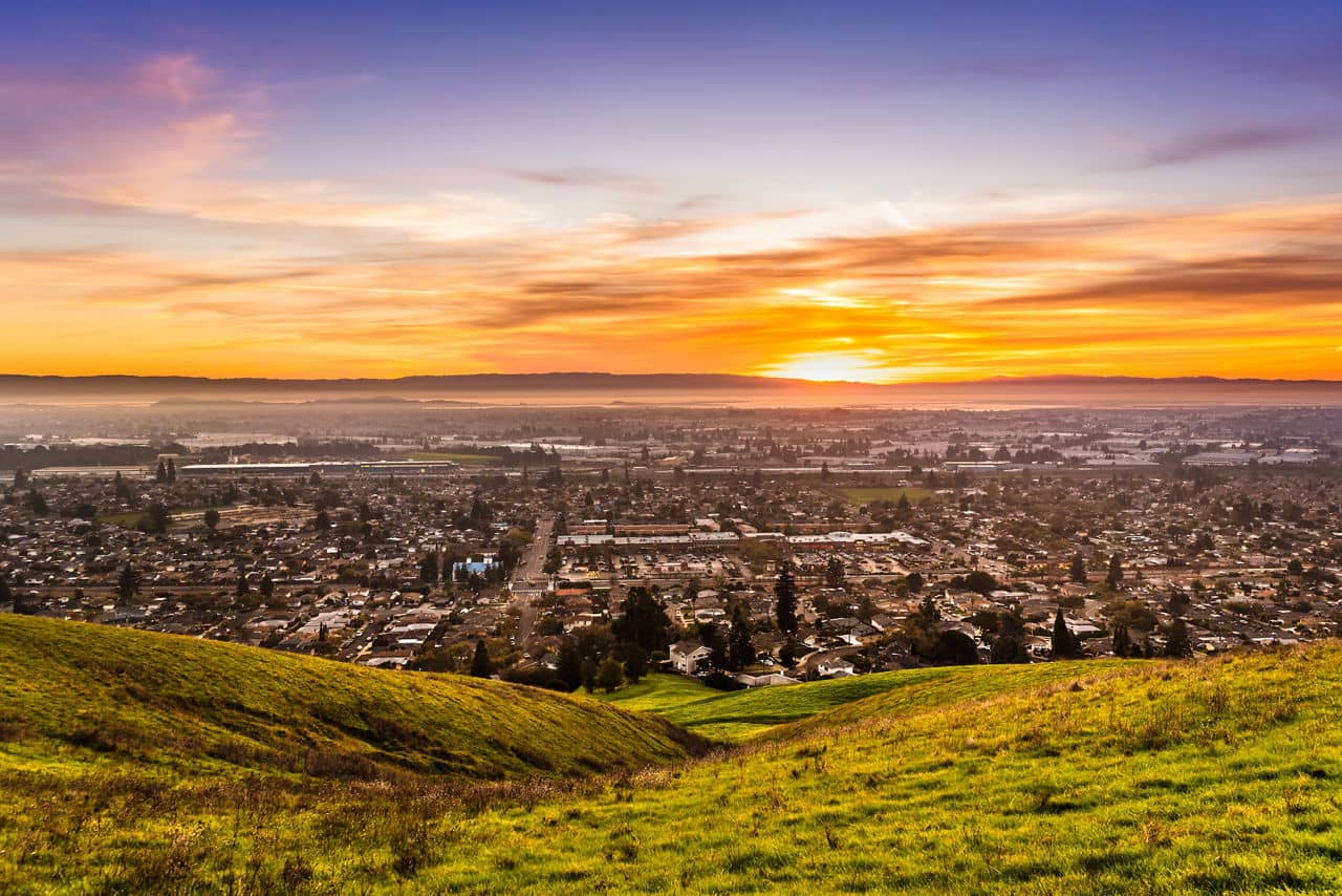 Sunset view over Hayward and San Leandro, California, featuring a lush green hillside in the foreground and a sprawling urban landscape.