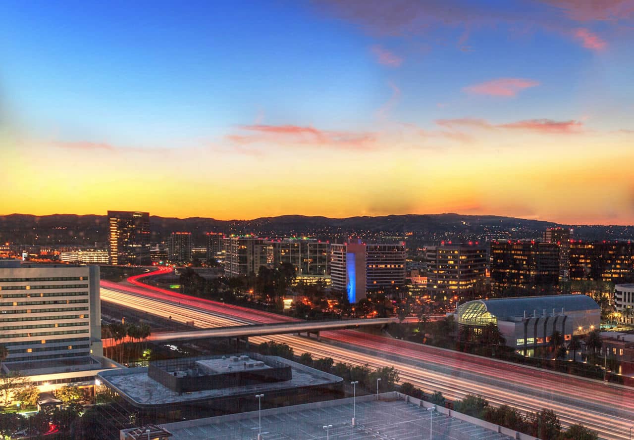 A vibrant evening view of Irvine, California, with illuminated buildings, a busy highway, and a colorful sunset in the background.