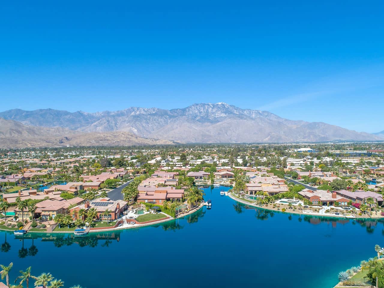 A scenic view of Rancho Santa Margarita, California, featuring a lake, trees, and houses surrounded by hills on a clear, sunny day.