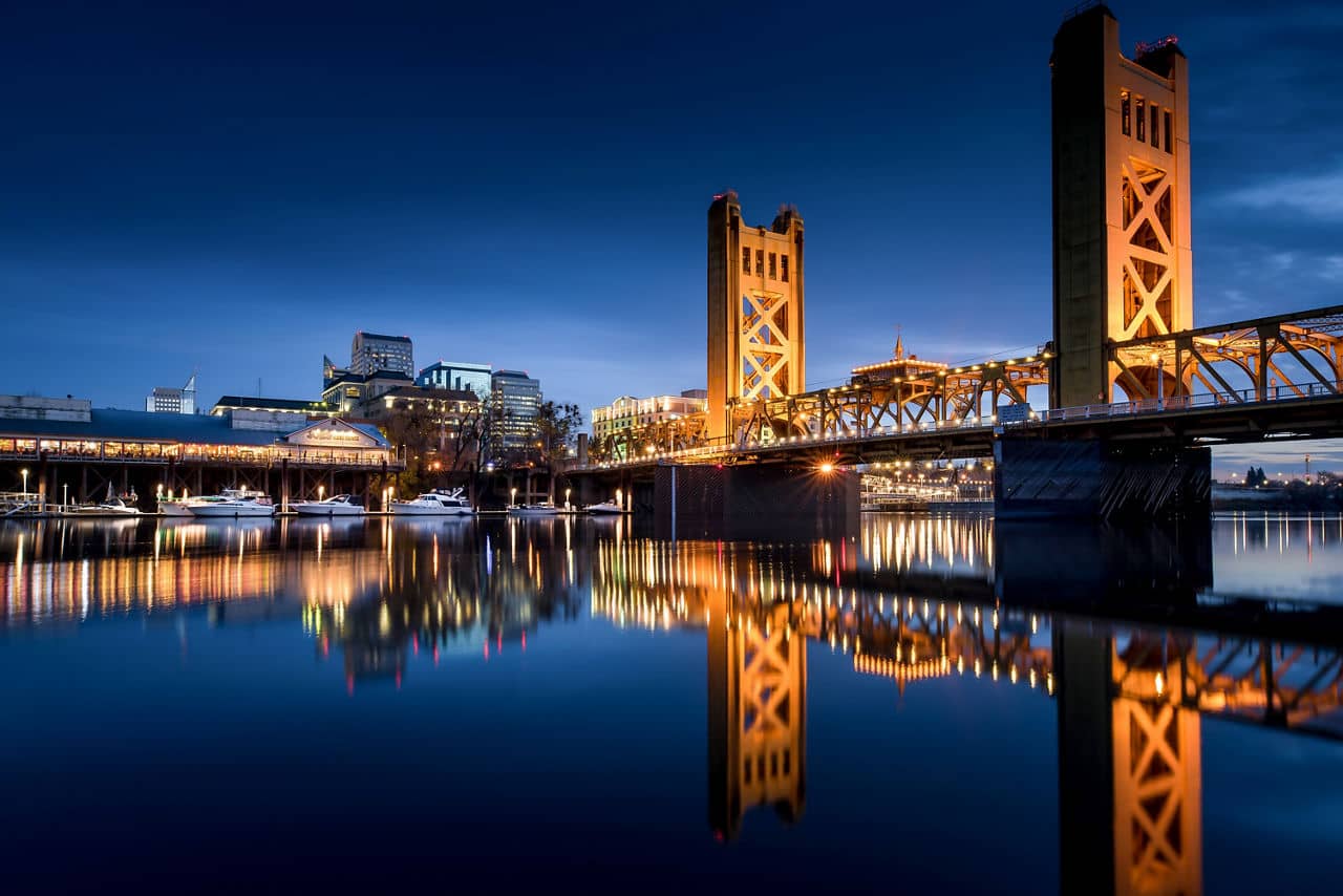 Tower Bridge and city skyline of West Sacramento, California, lit up at night with reflections in calm river water.