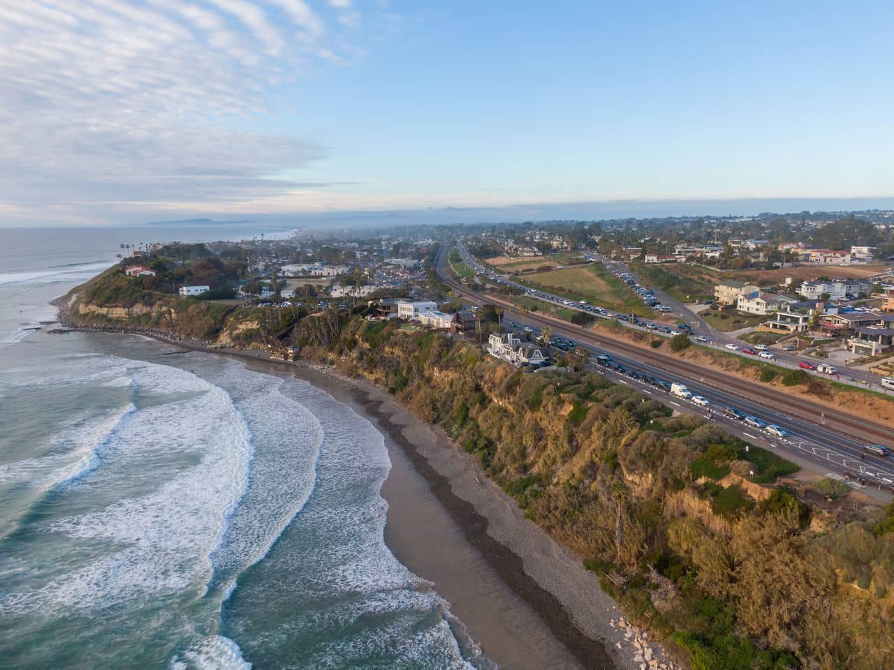 Aerial view of Encinitas, California, showcasing coastal cliffs, a sandy beach, waves, and a nearby highway with homes in the distance.