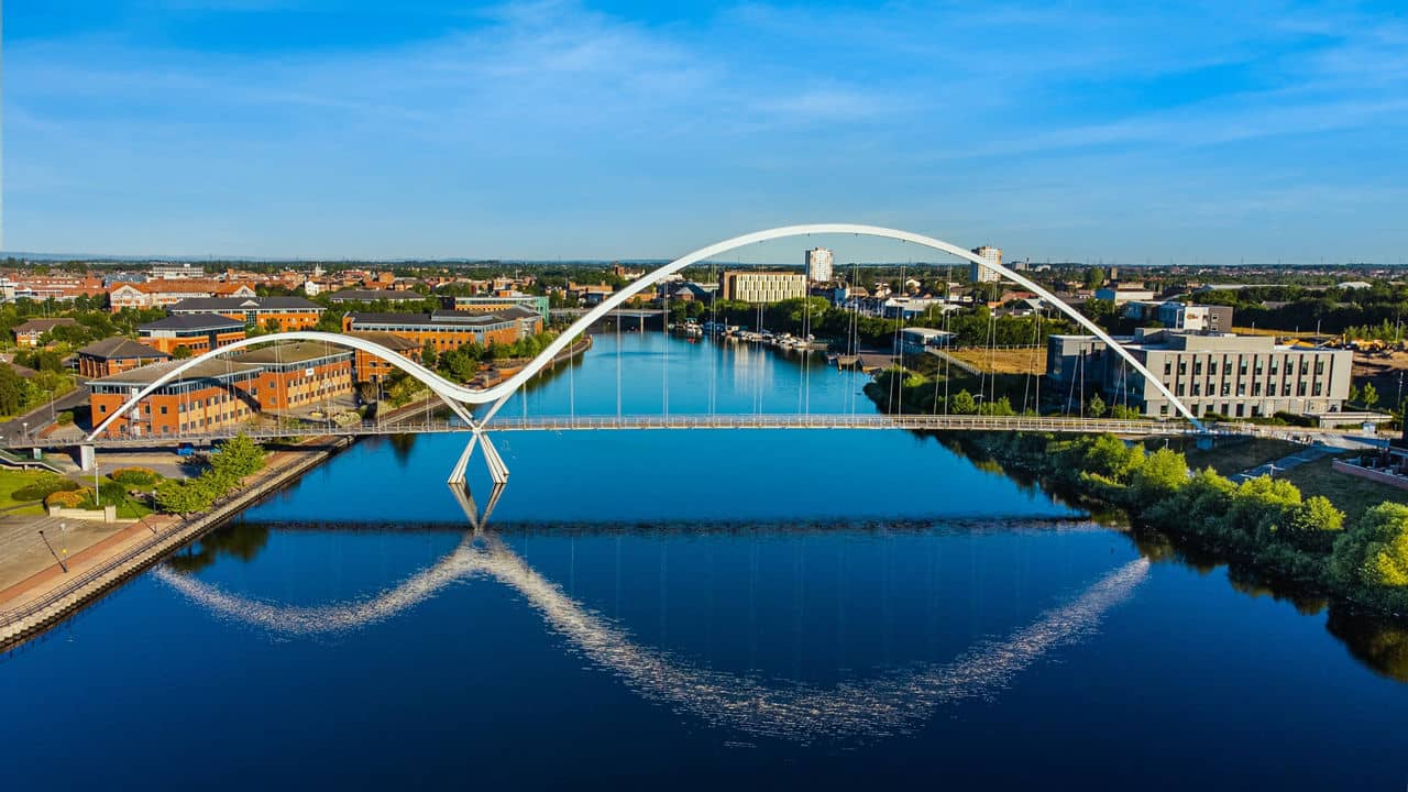 A modern arched bridge reflecting in a calm river, surrounded by buildings and greenery in Stockton, California, under a clear blue sky.