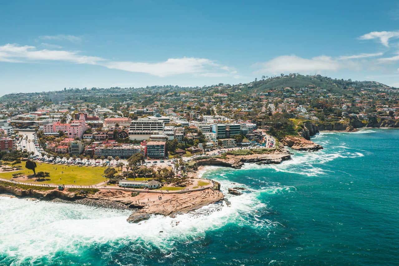 Aerial view of La Jolla, California, with turquoise waves crashing against rocky cliffs, a park, and hillside buildings.