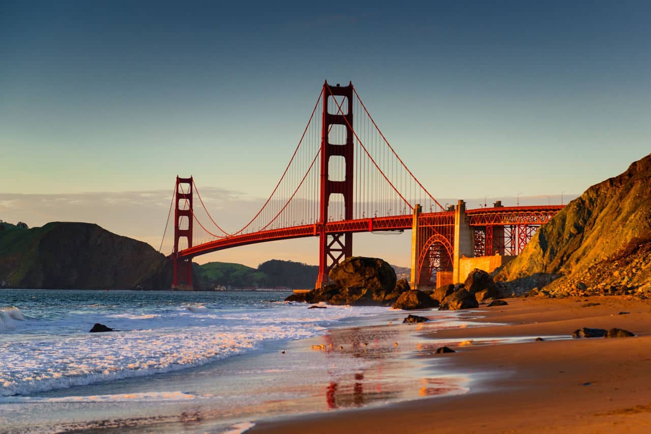 View of the Golden Gate Bridge from Marshall&apos;s Beach near Treasure Island, California, at sunset with waves lapping the shore.