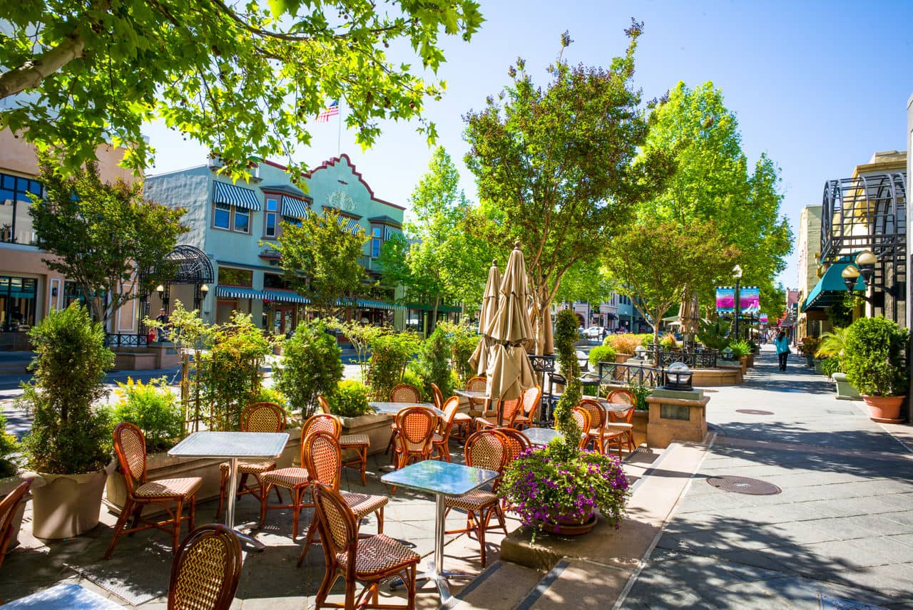 A sunny outdoor cafe seating area along a tree-lined shopping street in downtown Sunnyvale, California, with turquoise buildings and striped awnings.