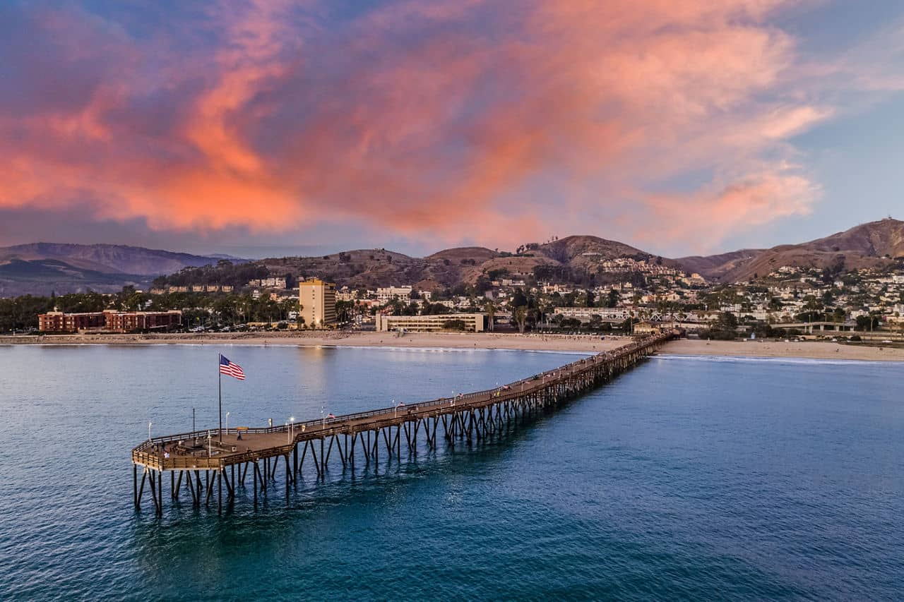 A scenic view of the Ventura Pier at sunset in Ventura, California, with hills, buildings, and a beach in the background.