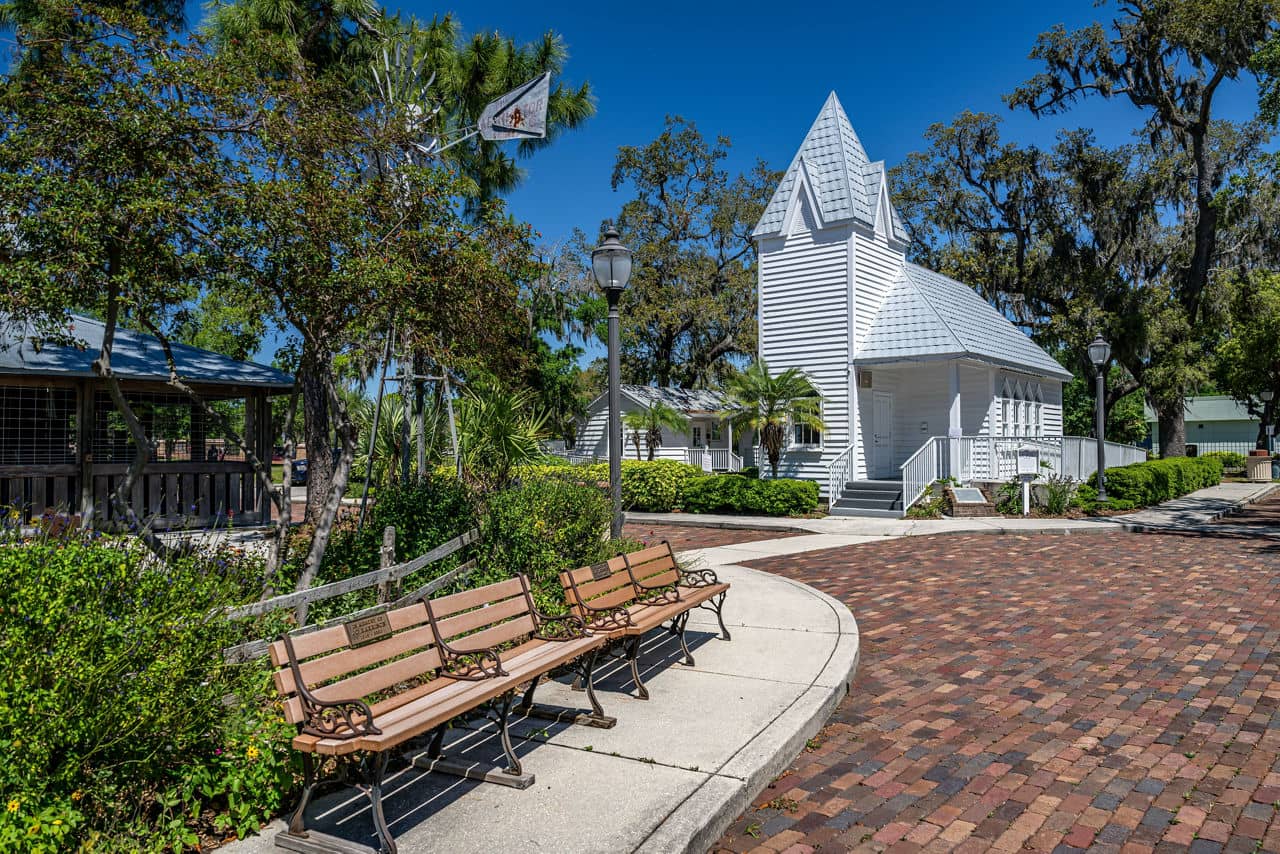 A quaint historic white church with a pointed roof in Palmetto, Florida, surrounded by greenery, brick paths, and wooden benches.