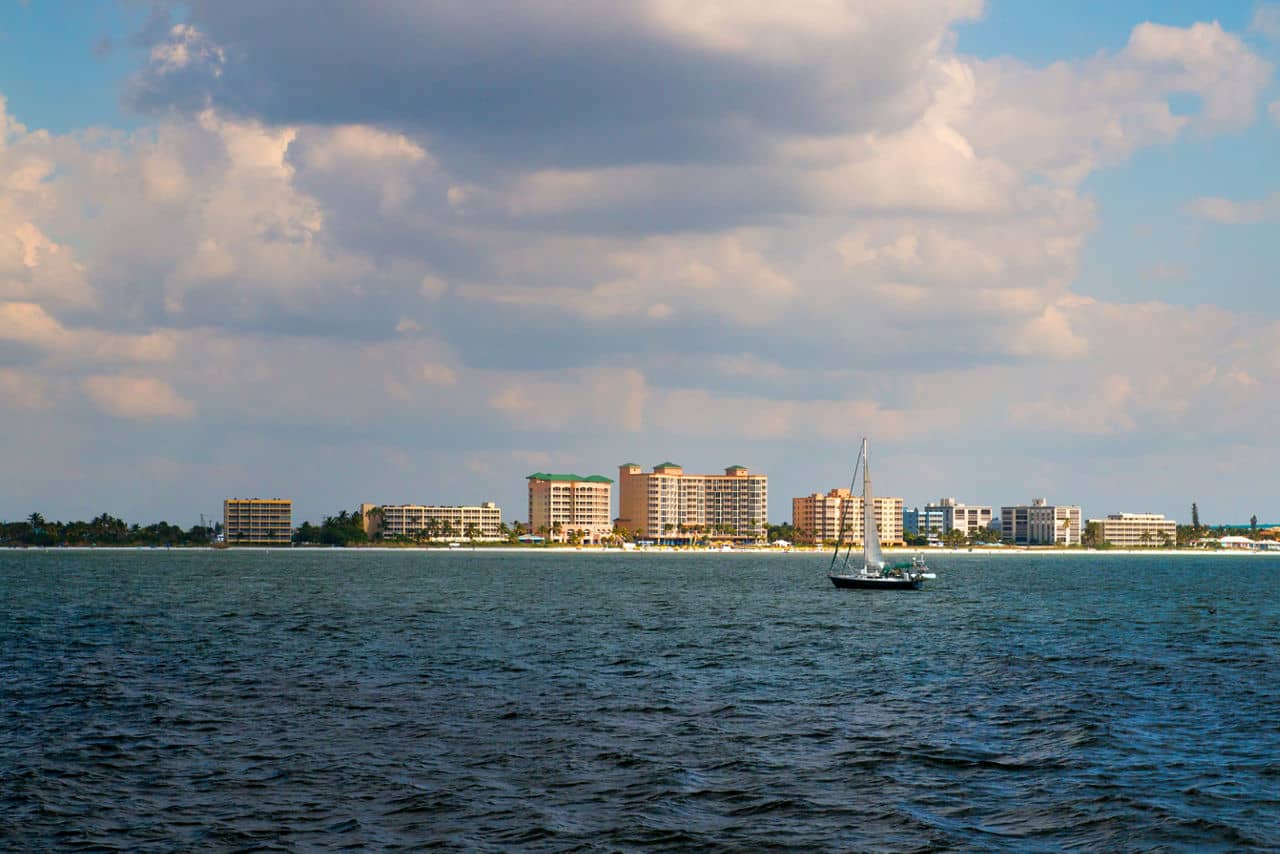 View of Estero, Florida&apos;s beachfront with a sailboat on dark blue water and beachside condos under puffy clouds.