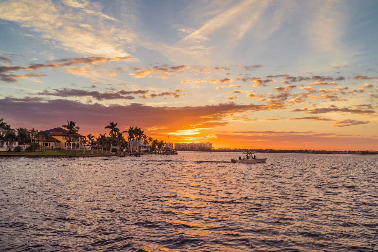 Stunning sunset over waterfront in Cape Coral, Florida, with a small boat cruising past waterfront homes with palm trees and colorful sky reflecting on the water.