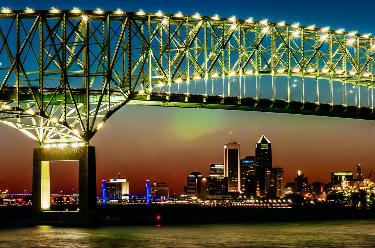 Night view of St. John&apos;s, Florida, with illuminated bridge spanning across the St. Johns River and Jacksonville city skyline glowing against an orange sunset sky.