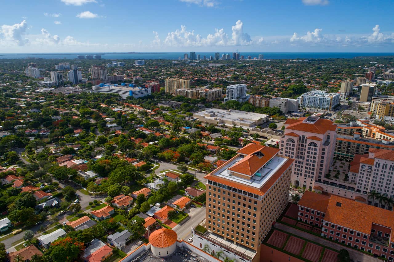  Aerial view of Coral Gables, Florida, with Mediterranean-style buildings featuring orange tile roofs and ocean visible in the distance.