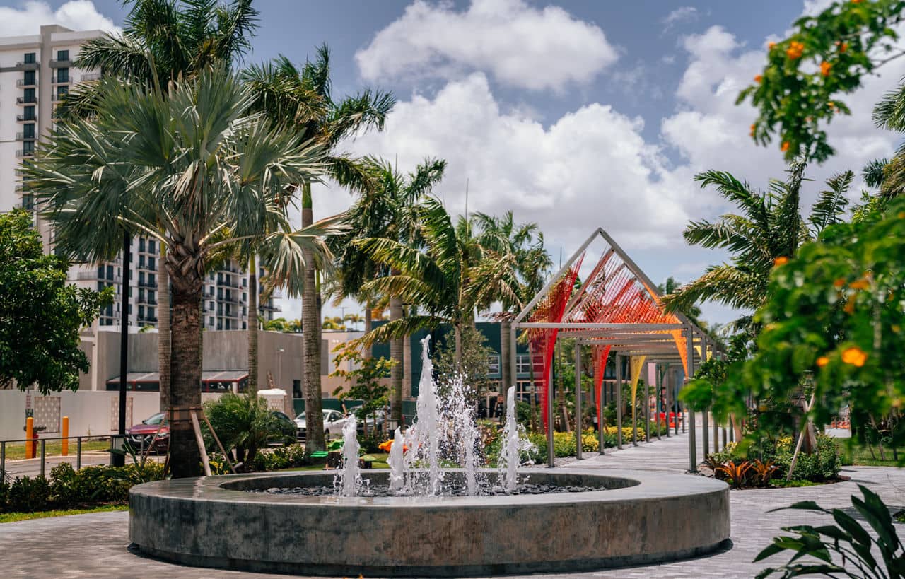 A modern park in Doral/Sweetwater, Florida, featuring a fountain, palm trees, and a colorful pavilion under a bright blue sky.