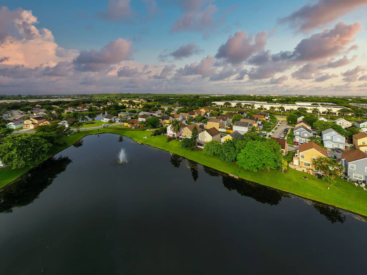 Aerial view of Miramar/Hialeah, Florida, showing colorful residential homes surrounding a retention pond with water fountain under pink clouds.
