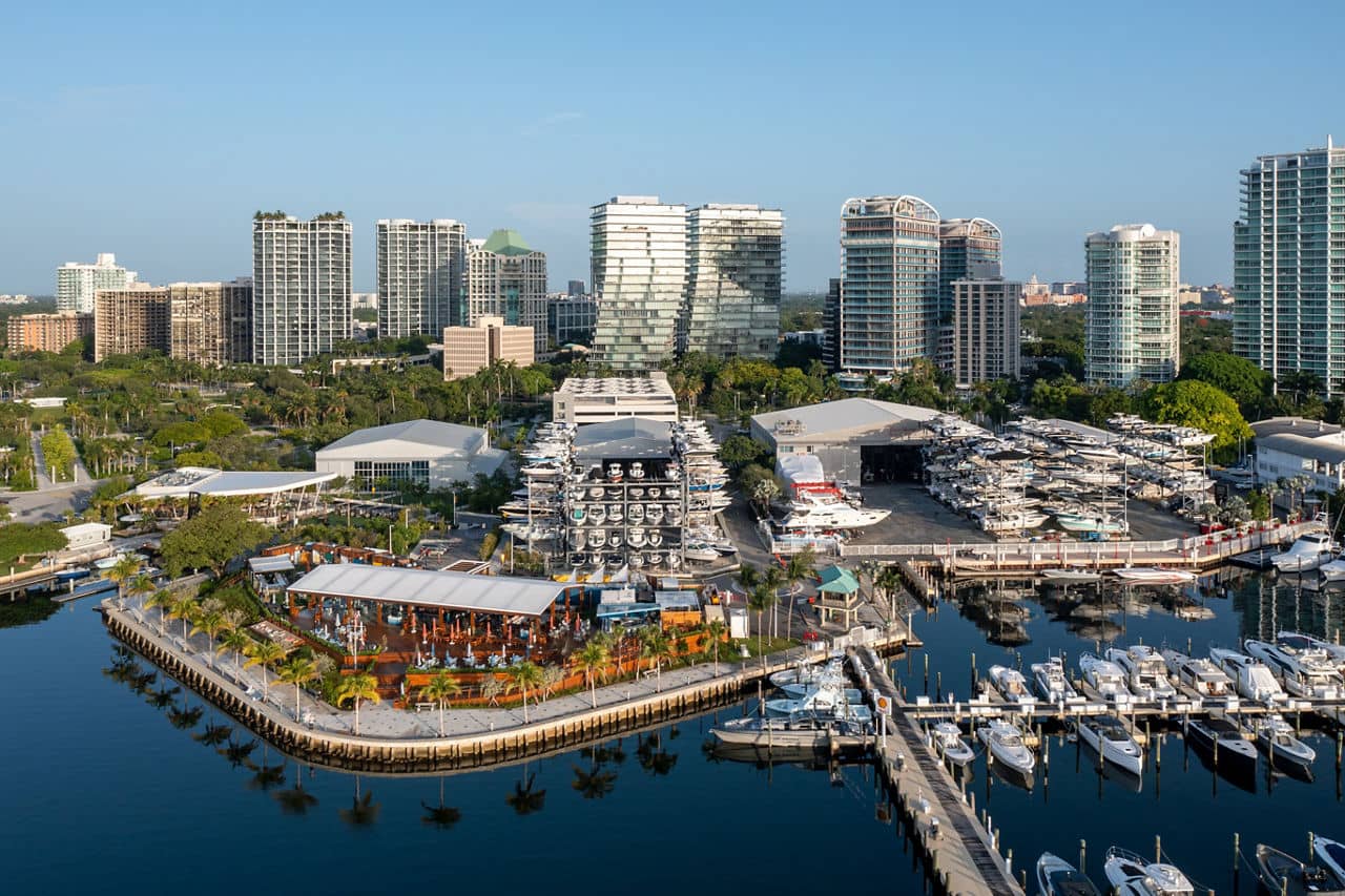 Aerial view of a marina in Miami, Florida, with luxury boats docked at piers, waterfront restaurants, and high-rise condominiums lining the skyline on a clear sunny day.
