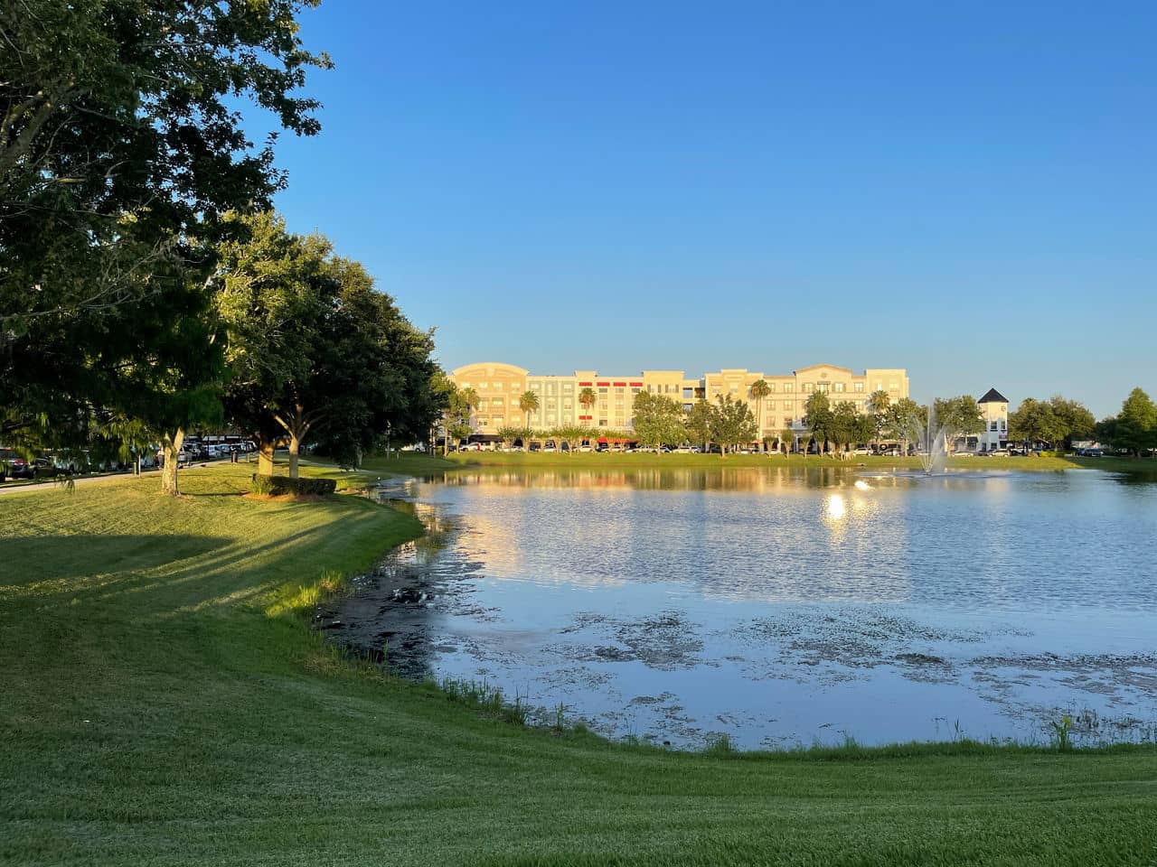 Lake view in Avalon Park, Florida, with apartment buildings across the water, green lawns, and a fountain in late afternoon light.