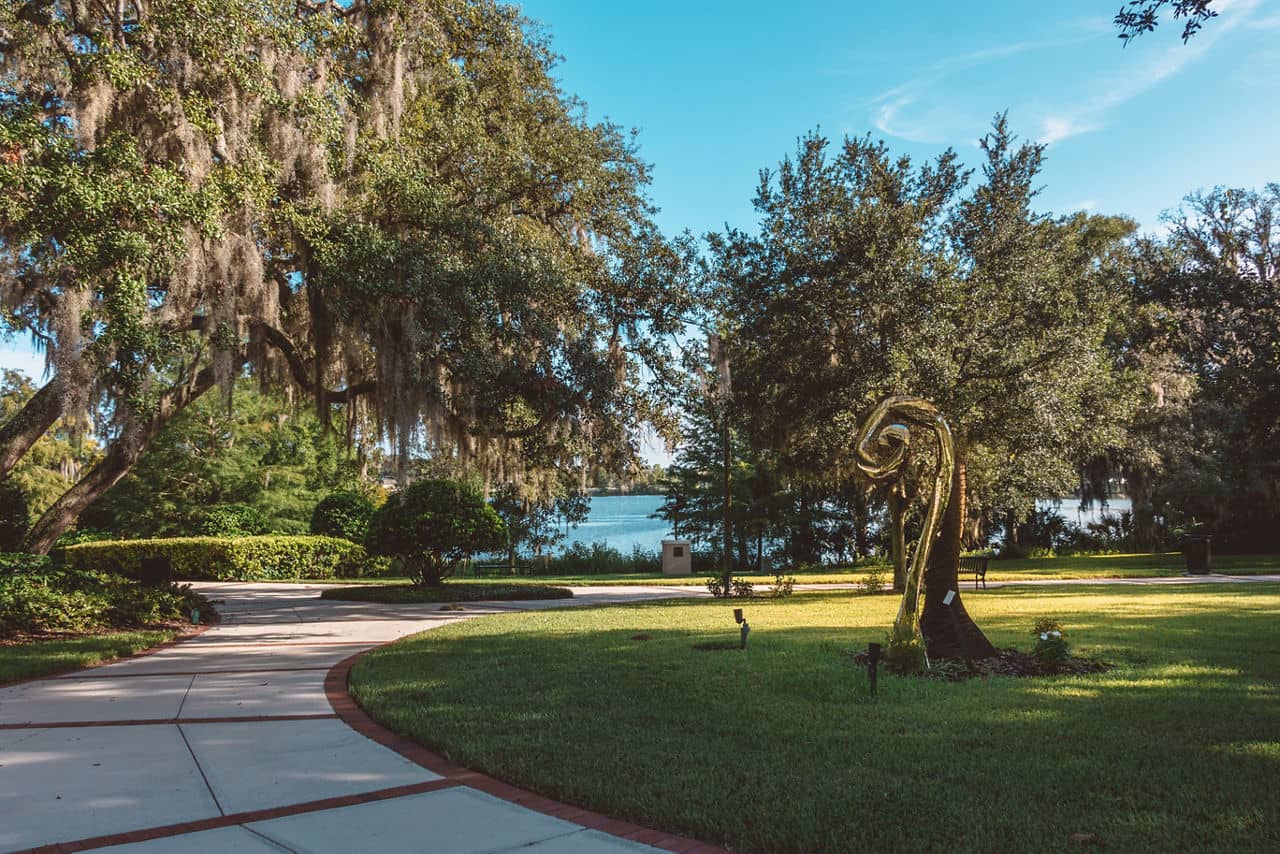 Park pathway in Seminole County, Florida, with Spanish moss hanging from oak trees, lawn sculpture, and blue lake in background.