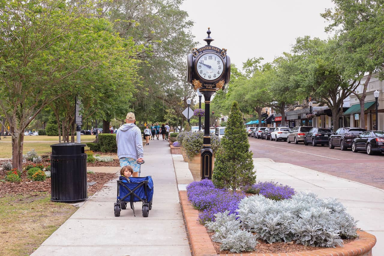 Downtown Winter Park, Florida&apos;s tree-lined sidewalk with decorative clock, purple flowers, and people walking past shops.