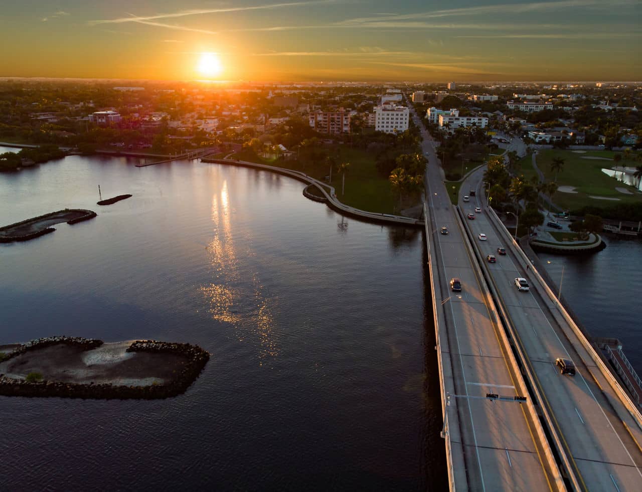  Aerial view of Lake Worth, Florida at sunset with bridge spanning across water, sun reflecting on lake, and city buildings.