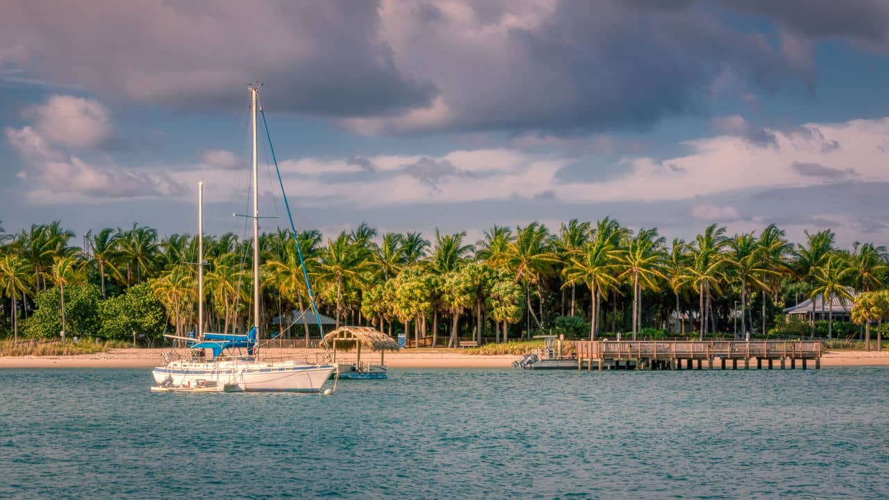 Sailboat anchored in turquoise waters near Palm Beach Gardens, Florida, with a wooden dock and palm tree-lined shore under a partly cloudy sky.