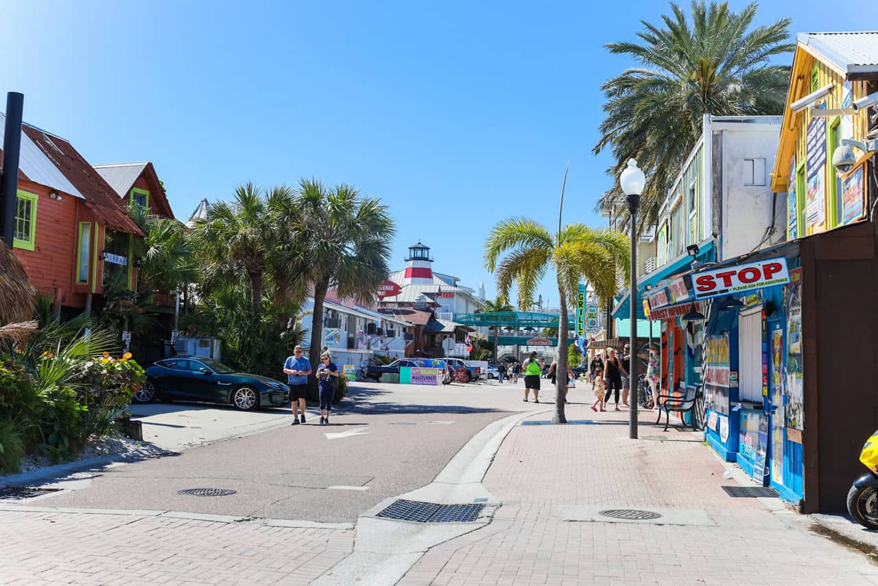 Colorful shopping street in Pinellas County, Florida, with lighthouse structure visible, palm trees, and tourists walking between shops.