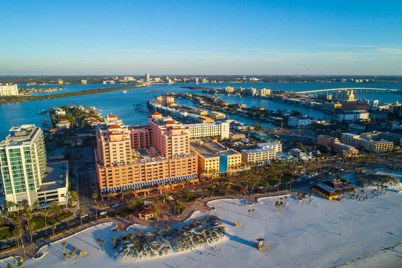 Aerial view of Clearwater, Florida, with white sand beach, pink hotel buildings, palm-lined streets, and blue waterways.