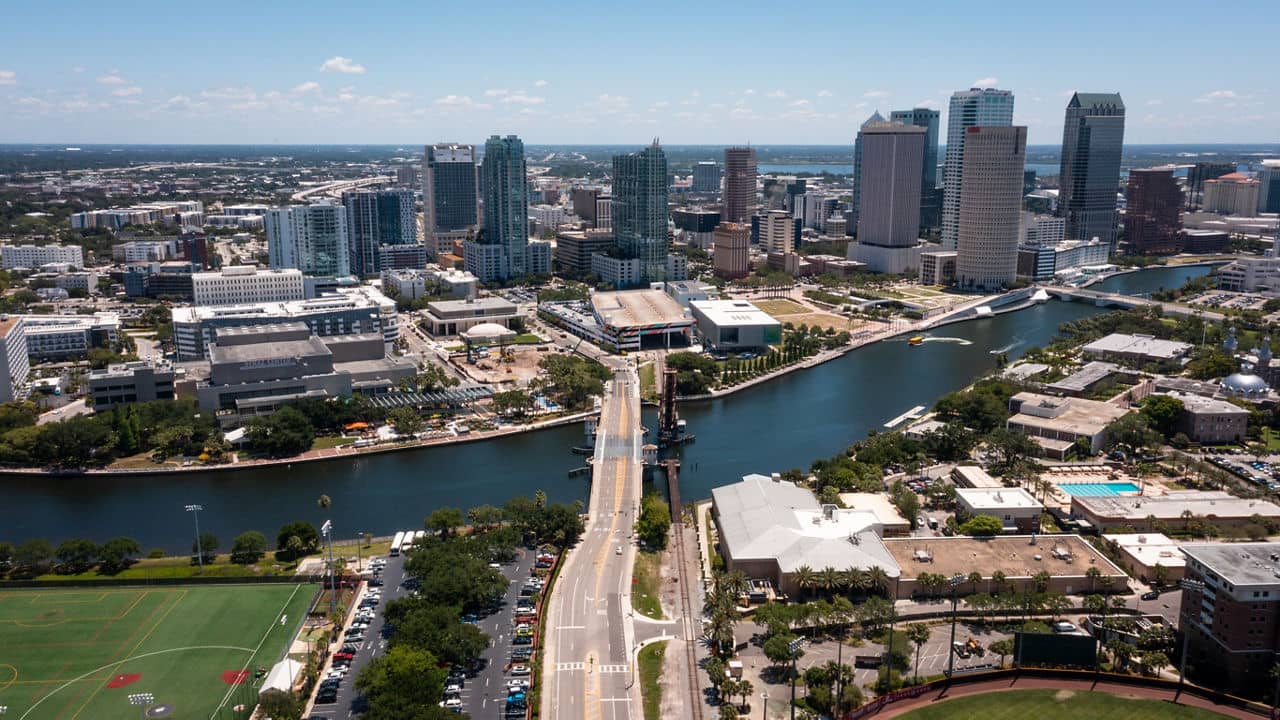 Aerial view of downtown West Tampa, Florida, showing the Hillsborough River cutting through the city with a bridge connecting West Tampa to the skyline of glass skyscrapers on a clear blue day.