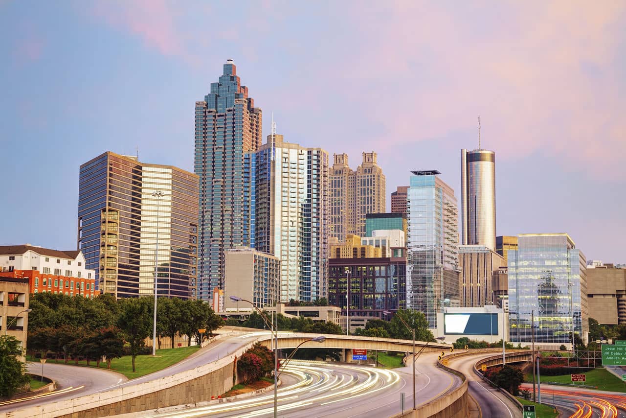 View from Norcross/Peachtree Corners, GA looking toward the Atlanta skyline with glass skyscrapers against a sunset sky.
