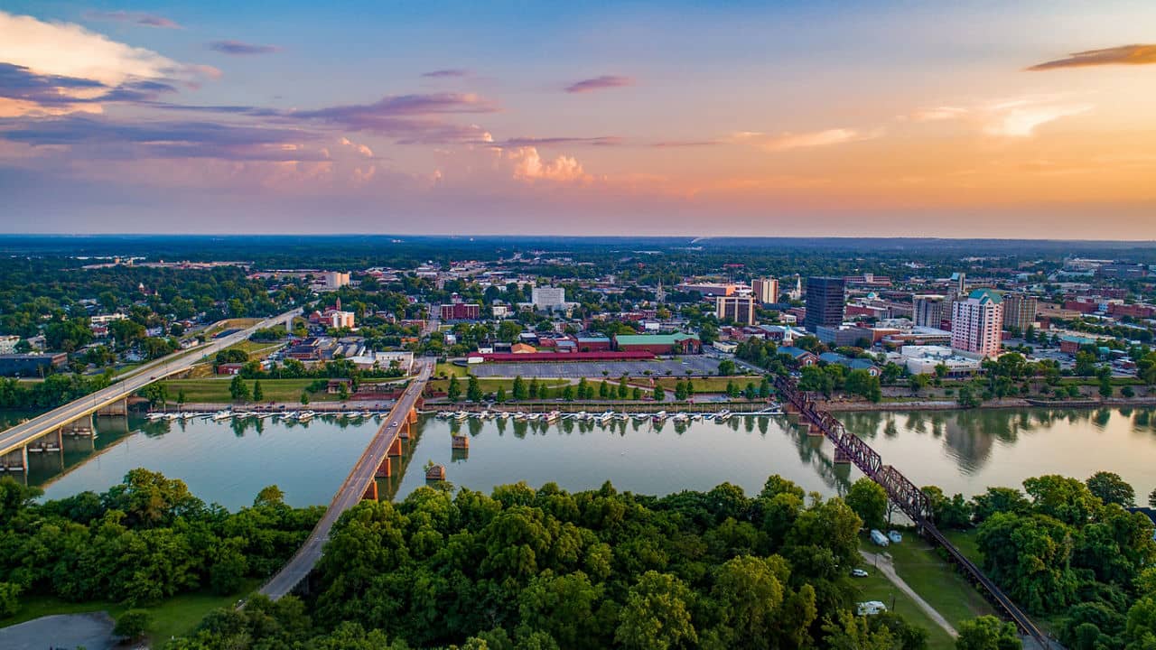 Aerial view of Augusta, GA at sunset showing the Savannah River with bridges, downtown buildings, and tree-lined shores.