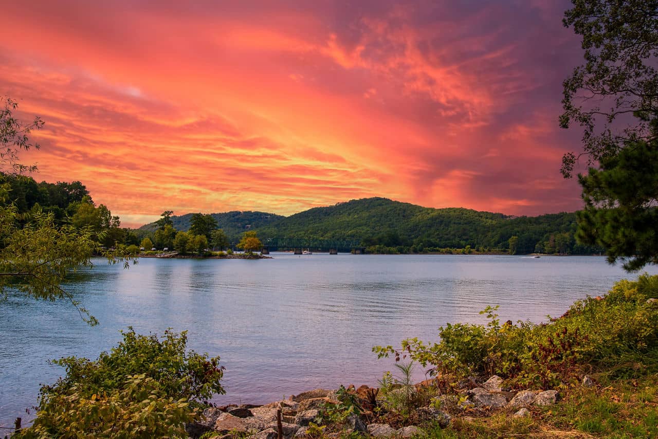 Acworth, GA lake at sunset with orange-pink sky, green mountains in the background, and calm water surrounded by trees.