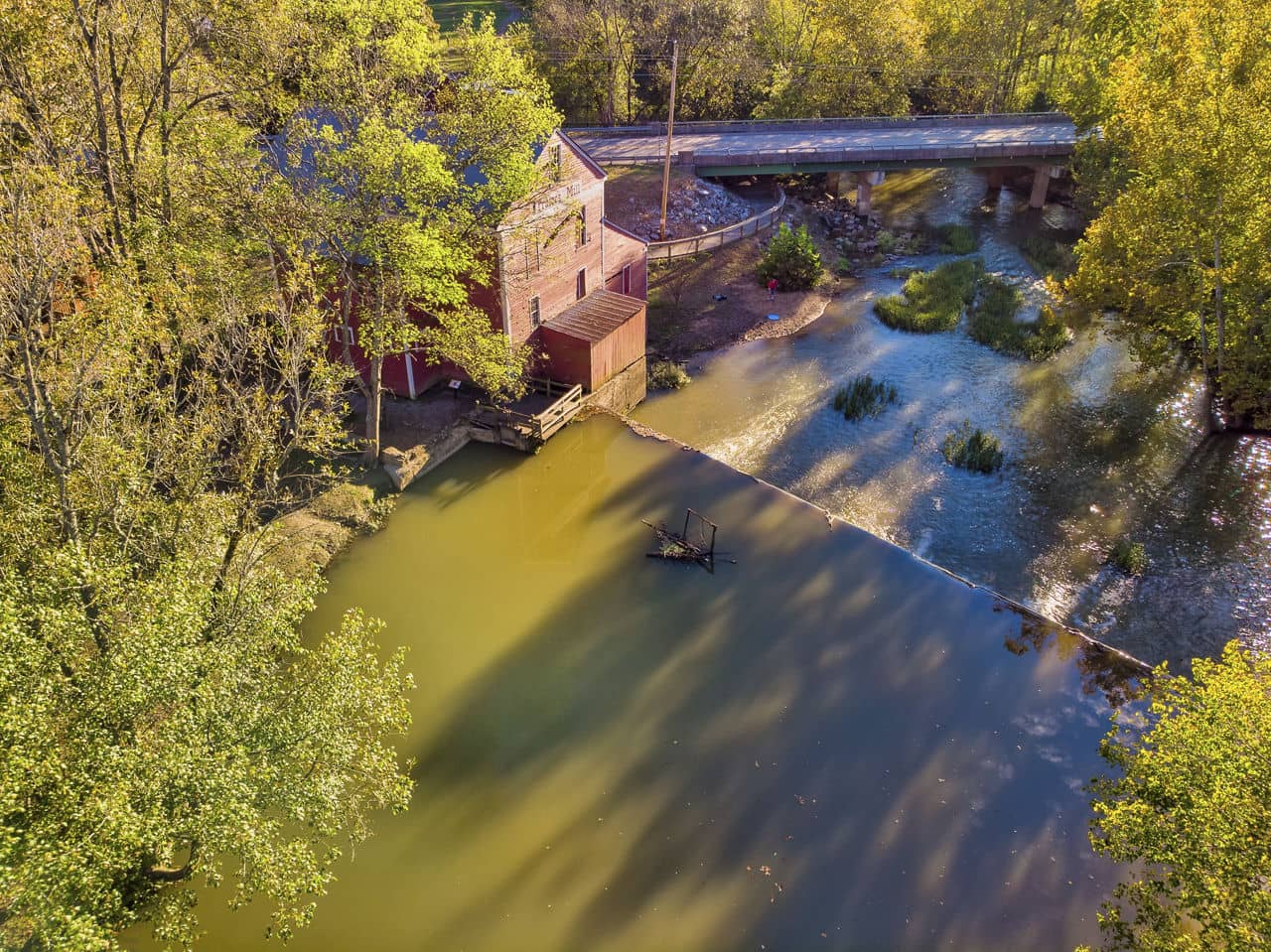 View of historic mill building beside creek in Dalton, GA with small dam creating a pond, surrounded by autumn trees.