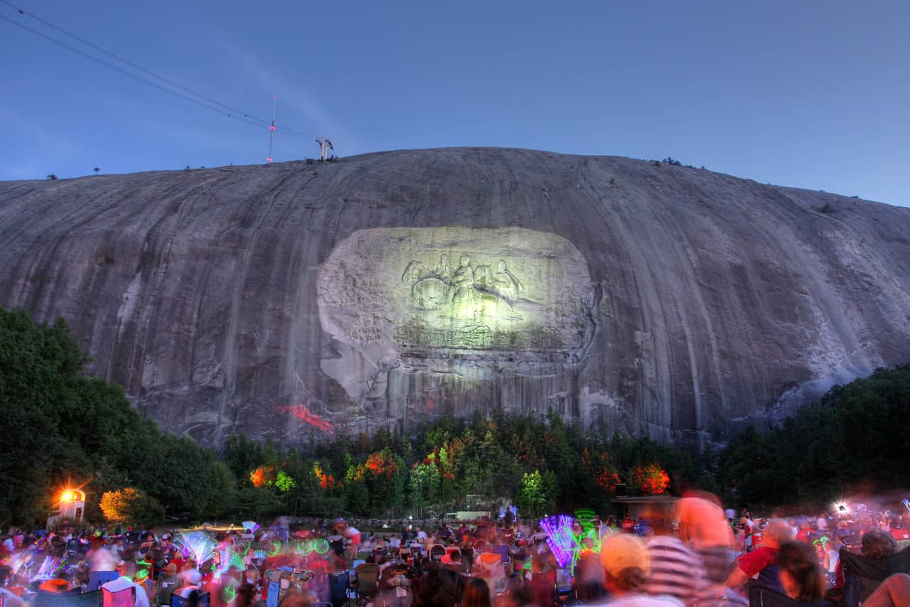 Illuminated carving on Stone Mountain, GA with nighttime crowd below holding colorful glowsticks during a special event.