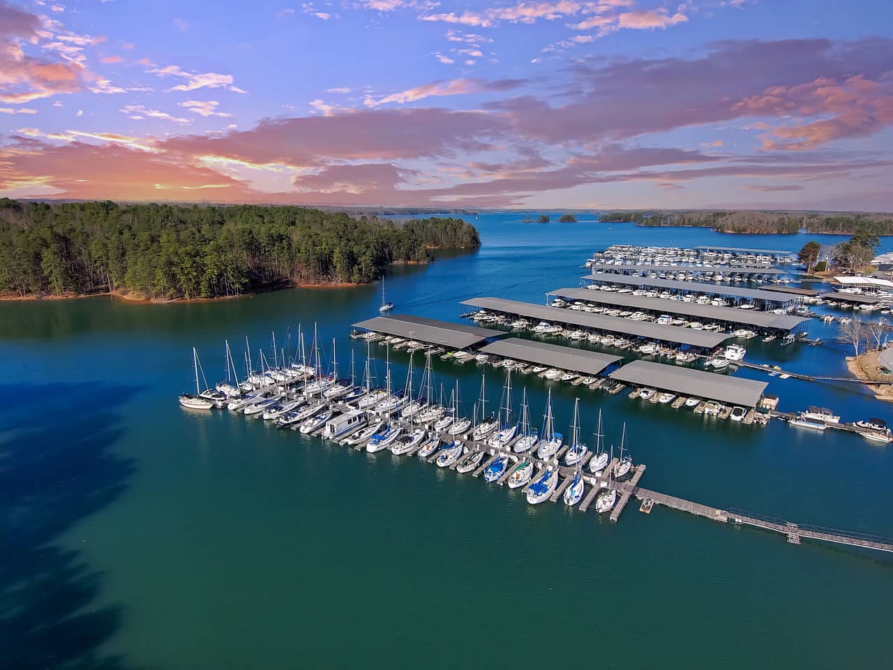 View of a marina in Buford/Suwannee, GA with rows of white sailboats and boat docks on a lake surrounded by pine trees.