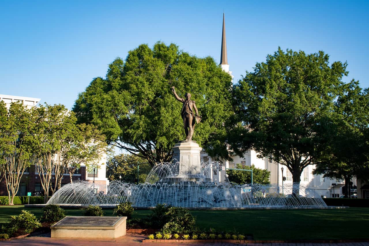 Fountain plaza in Lagrange/Newnan, GA with a statue of a historical figure, water jets, and a church steeple beyond trees.