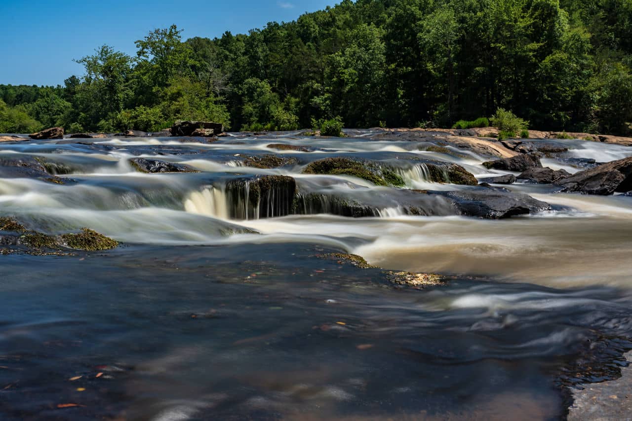 Flowing rapids over dark rocks at a river in Rockdale County, GA with a green forest backdrop and clear blue sky above.