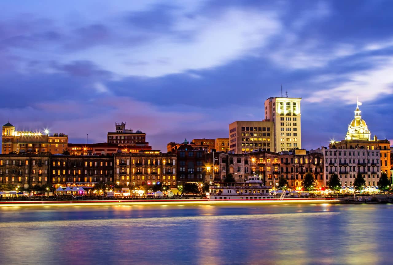 Evening waterfront view of historic Savannah, GA with illuminated buildings, gold-domed city hall, and the river.