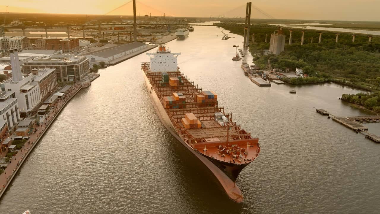 A large, loaded cargo ship sailing down the Savannah River in Port Wentworth, GA with a suspension bridge at sunset.
