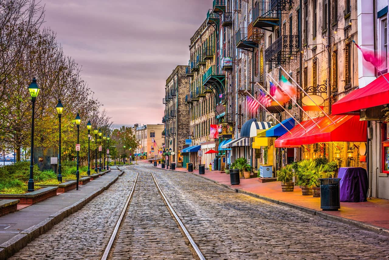 Historic cobblestone street in Savannah, GA with trolley tracks, colorful storefronts, and wrought-iron balconies at dusk.