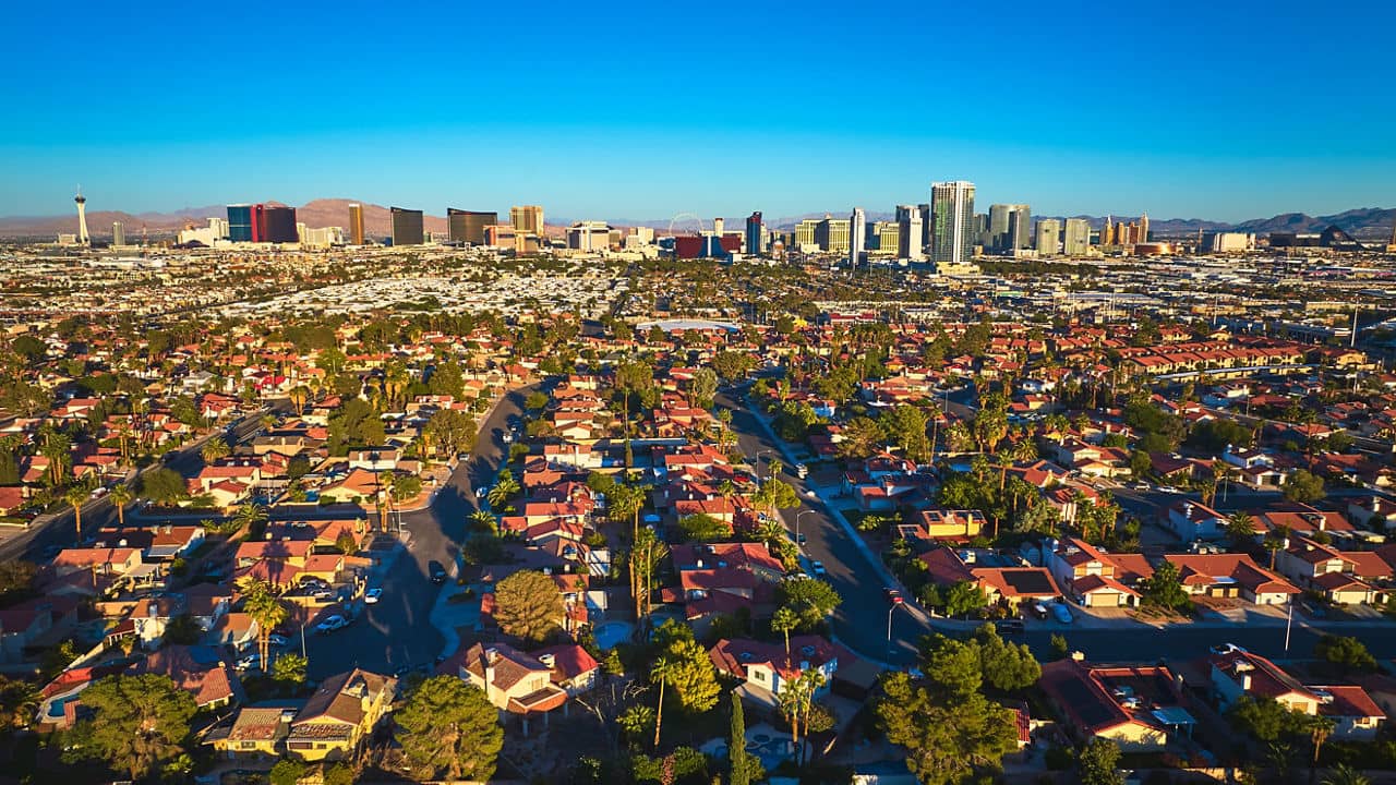 Aerial view of suburban neighborhoods in Las Vegas Nevada with The Strip skyline and desert mountains in the distance. Warm golden hour sunlight highlights the dynamic blend of city and residential life.