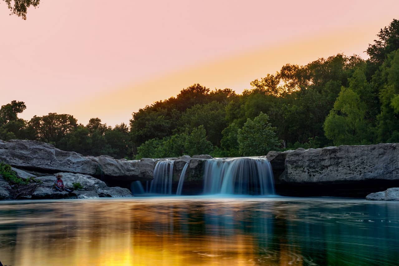 A peaceful waterfall in Bluff Springs/Onion Creek, Texas, surrounded by lush greenery under a colorful sky at sunset.