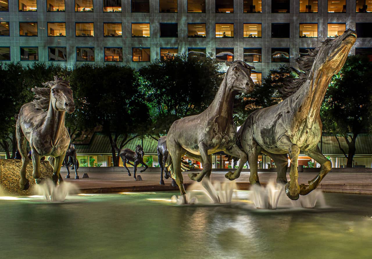 Three bronze horse statues are splashing in water at night in Irving/Las Colinas, Texas. Buildings are visible in the background.